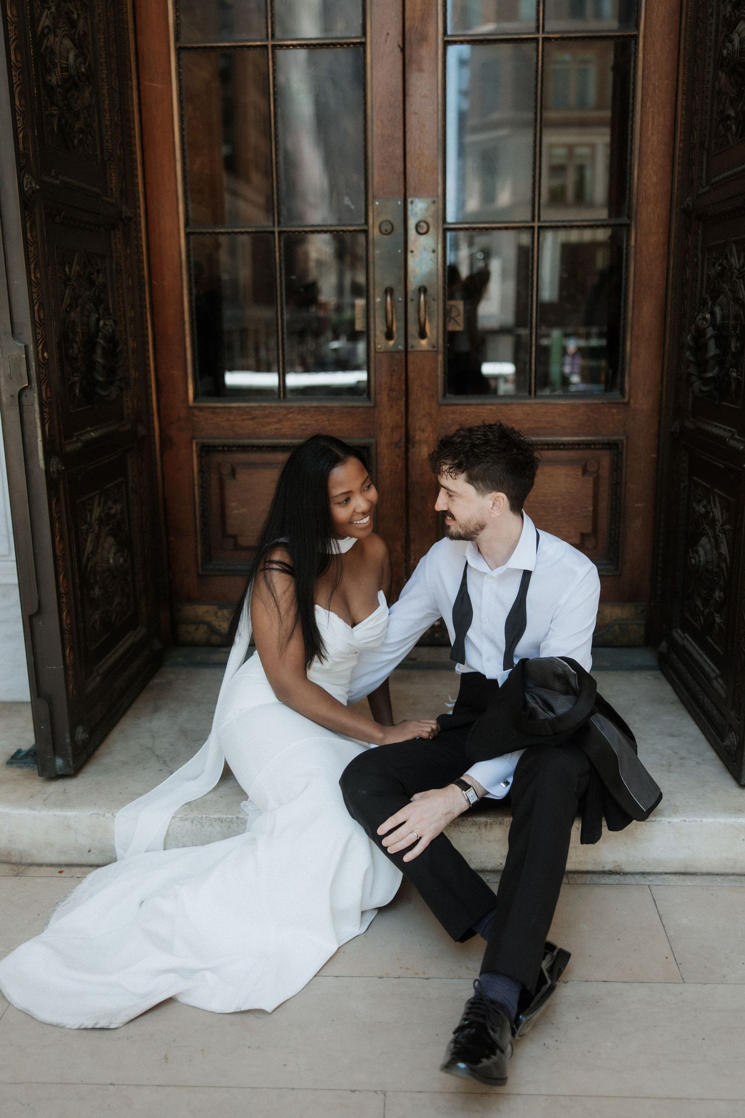Newlyweds share an intimate moment while sitting against wooden doors with glass panels.