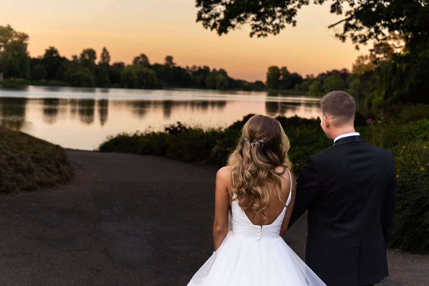 Couple in wedding attire walks hand in hand along a lakeside path at sunset creating a romantic silhouette | Hever Castle, Kent