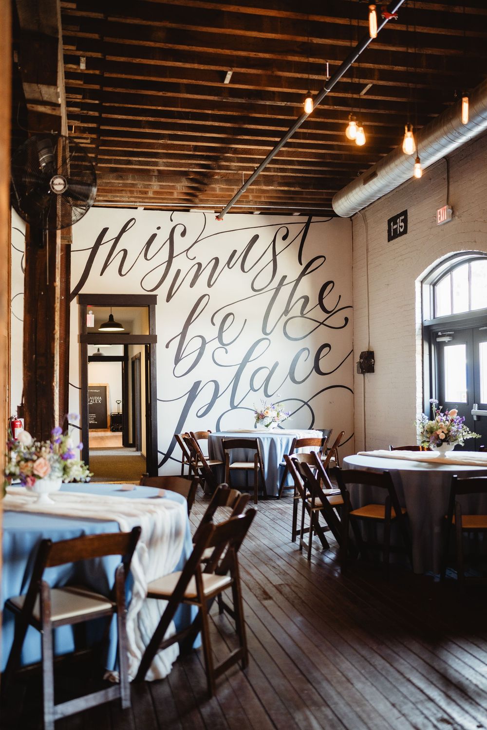 Rustic restaurant interior with wooden floors and a large decorative script text on the wall above dining tables.