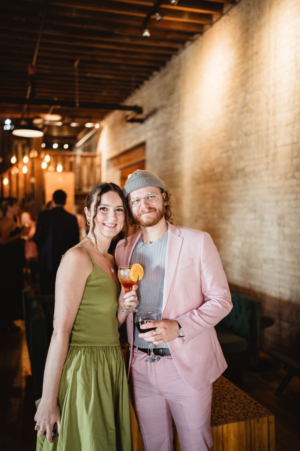 Couple at an indoor event venue with exposed brick walls, person in pink suit holding cocktail and companion in green dress.