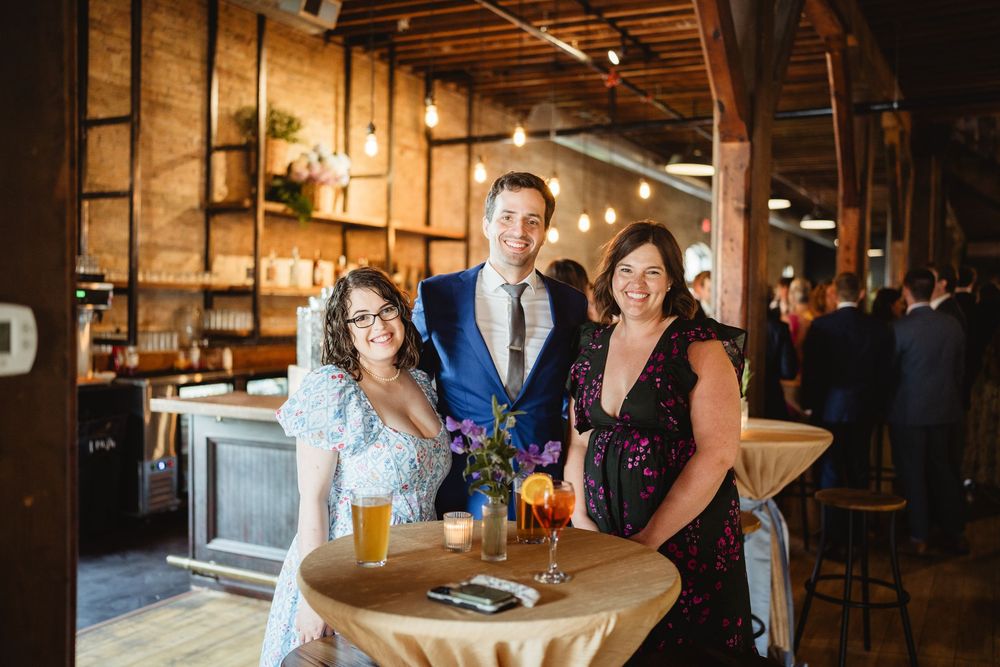 Three people posing together at a rustic event venue with cocktails and table decorations.