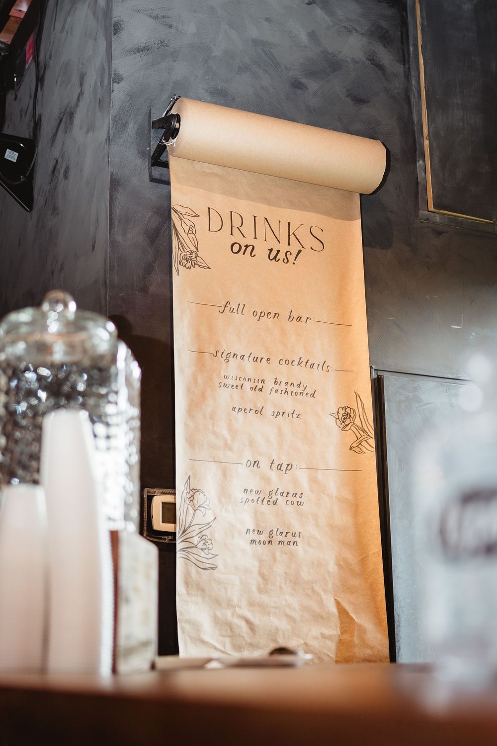 Vintage-style drinks menu written on brown kraft paper scroll mounted on dark wall.