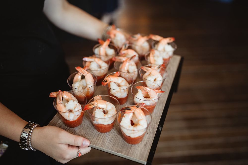 Hands reach for gourmet cupcakes topped with cream cheese frosting and garnished with red decorations arranged on a wooden board.