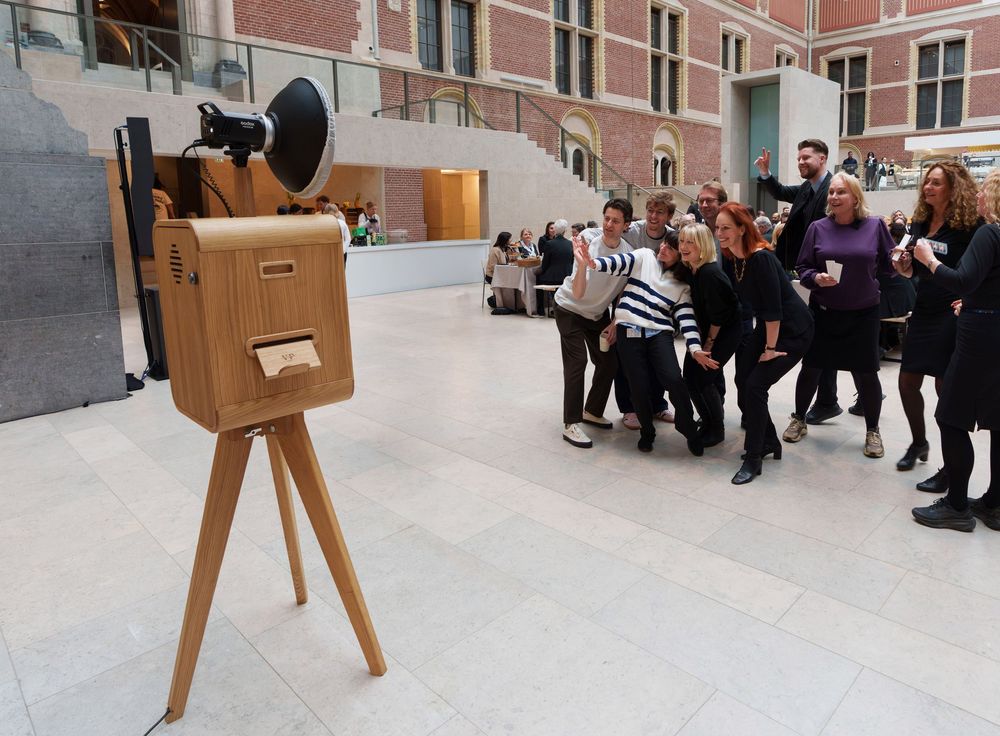 Group of people posing for a vintage wooden camera on a tripod inside a modern museum atrium.