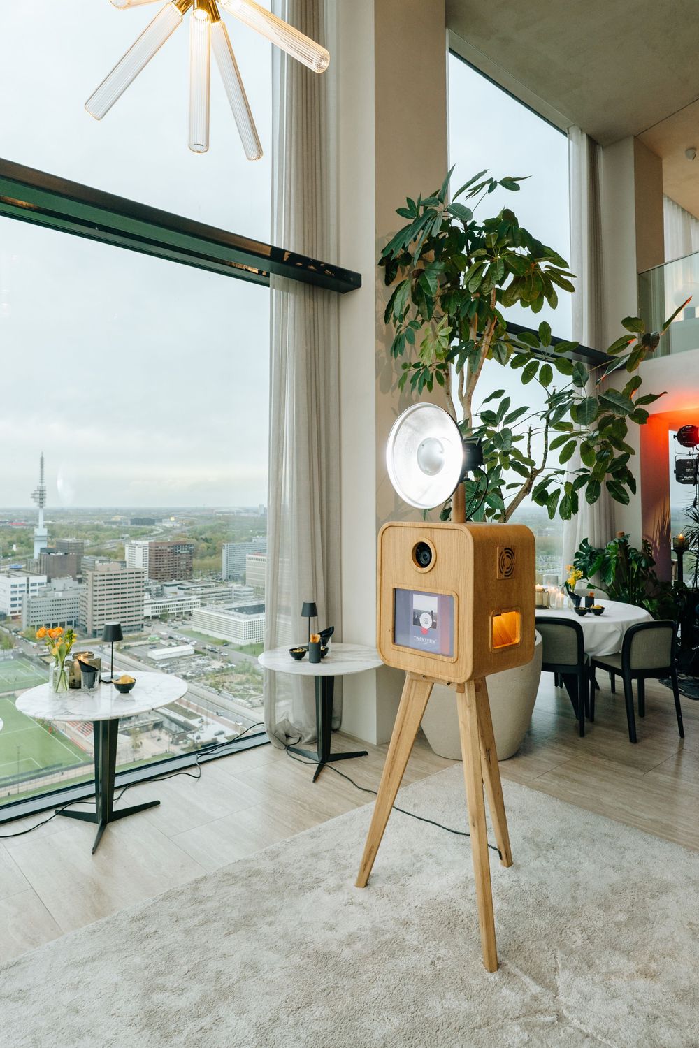Elegant wooden photo booth on tripod in modern high-rise event space with panoramic city views.
