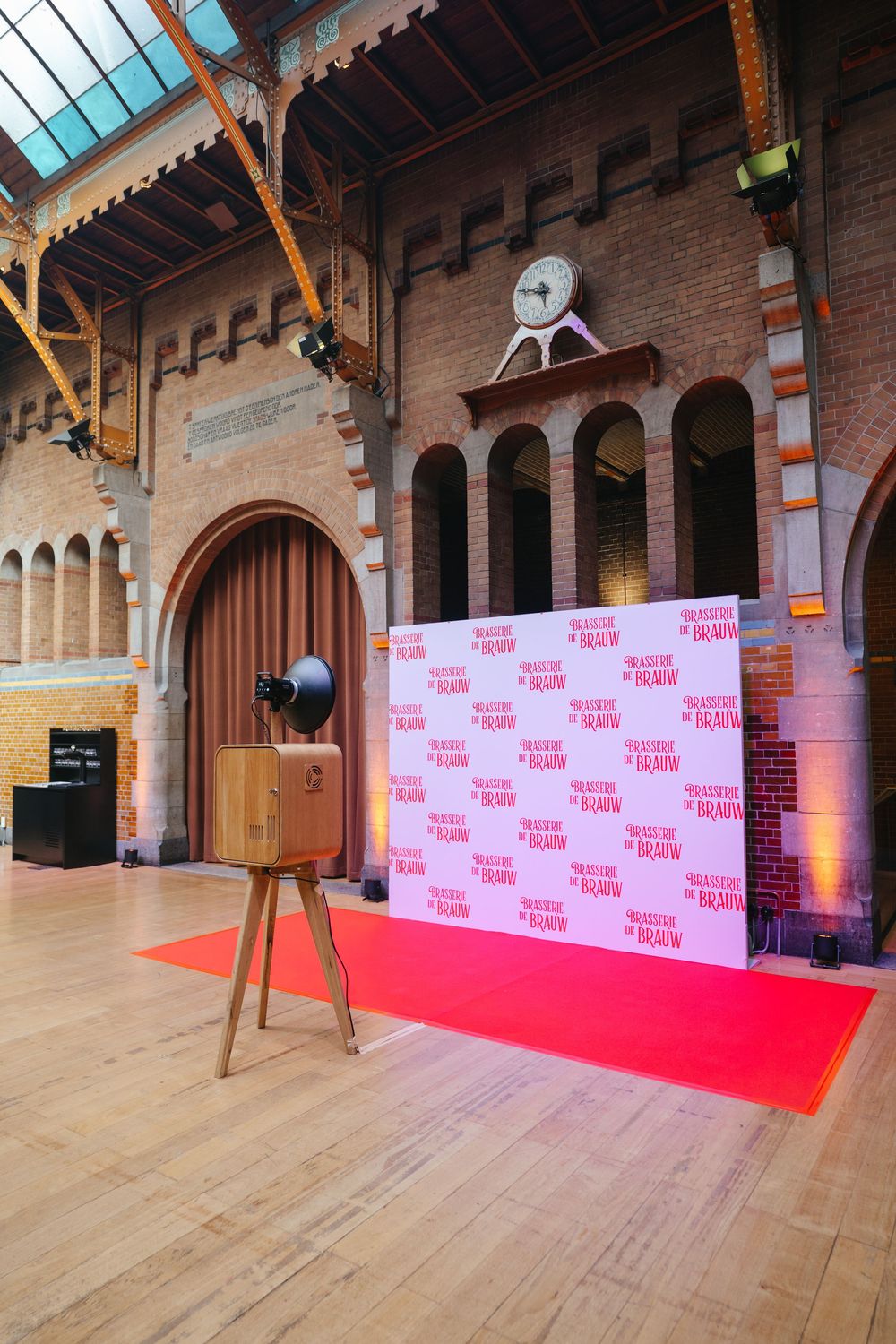 Vintage wooden camera photo booth on tripod beside red carpet step-and-repeat banner in historic hall.