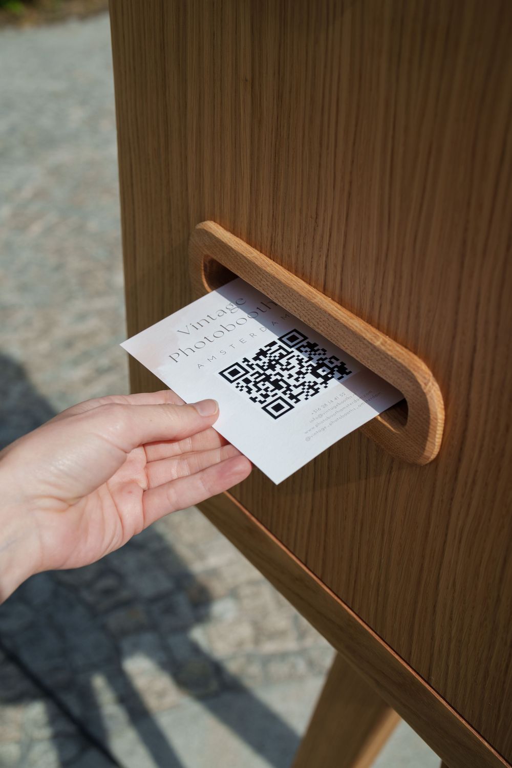 Hand inserting a business card with QR code into a wooden slot on a photo booth kiosk outdoors.