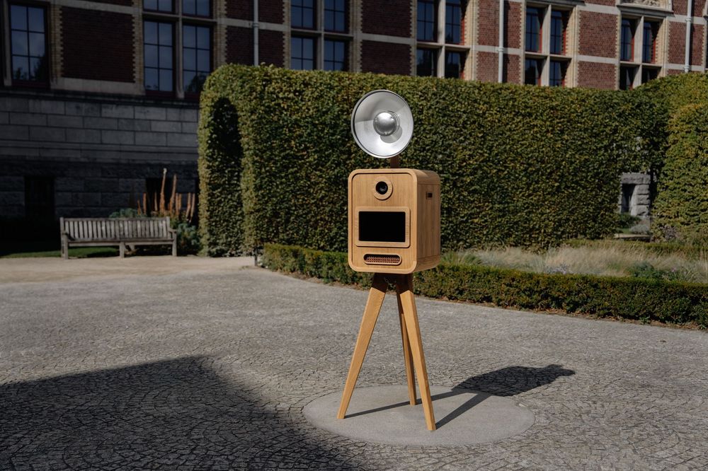 Classic wooden photo booth with studio flash on tripod placed on cobblestone courtyard beside historic building.