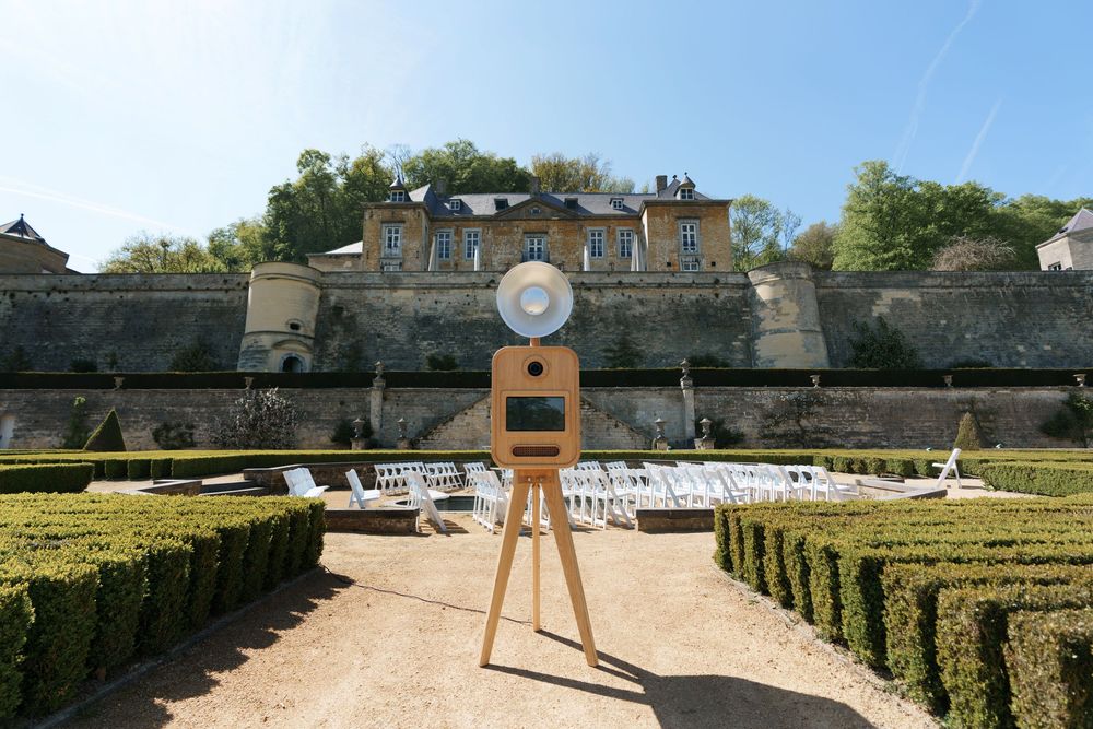Elegant photo booth on wooden tripod in formal garden with manicured hedges and historic chateau backdrop.