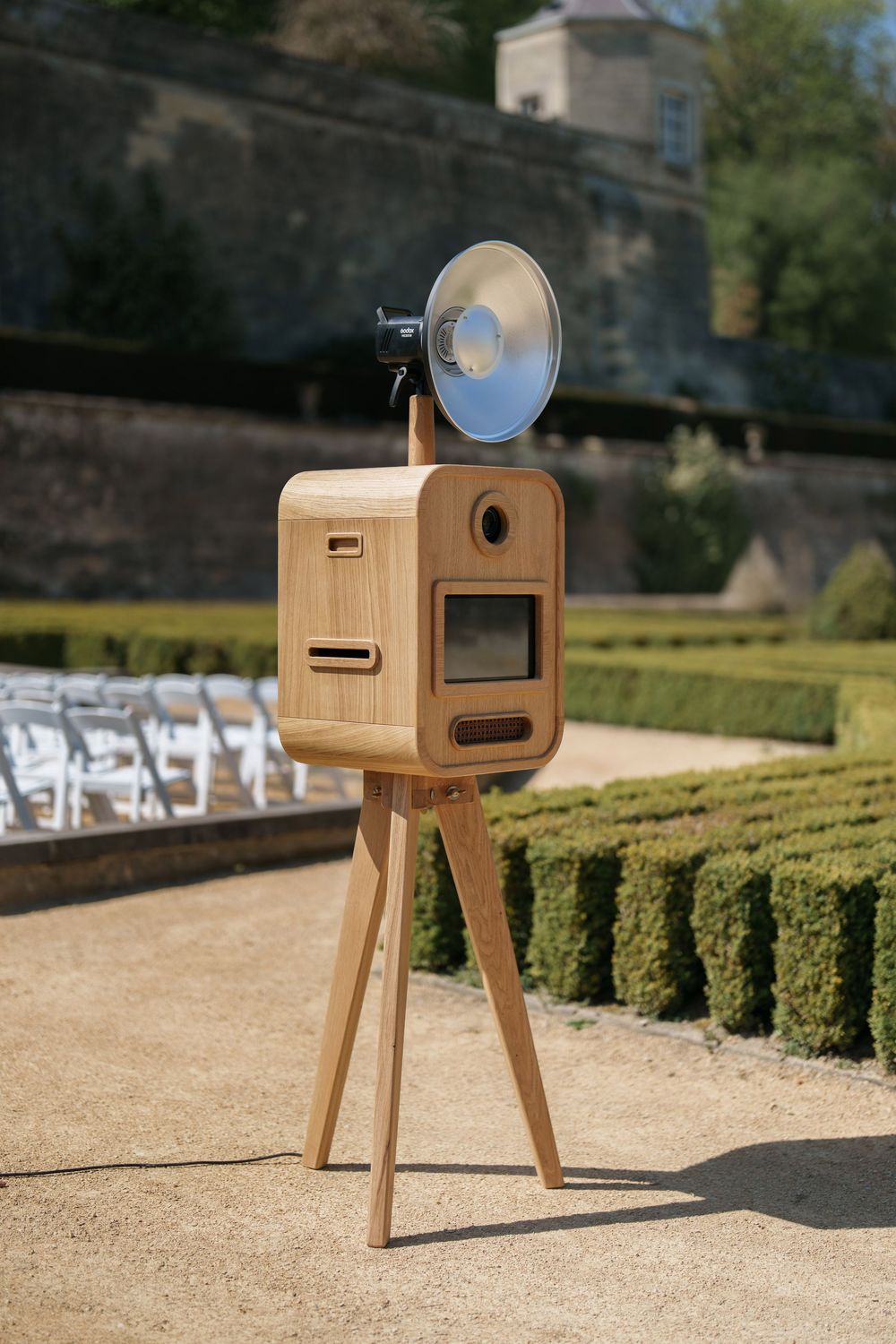 Vintage wooden photo booth on tripod with flash lamp in elegant garden setting with manicured hedges.