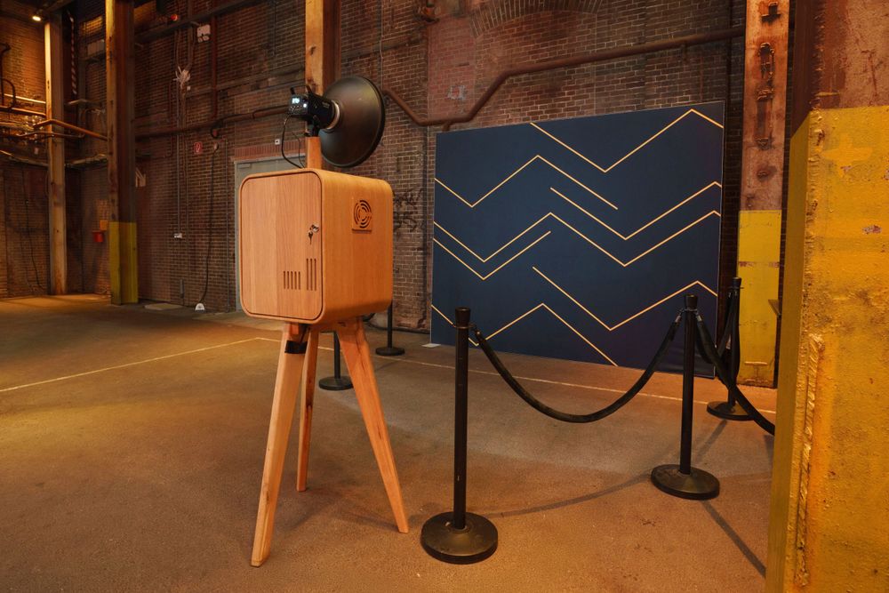Rustic wooden photo booth on tripod beside blue chevron backdrop in industrial warehouse venue.