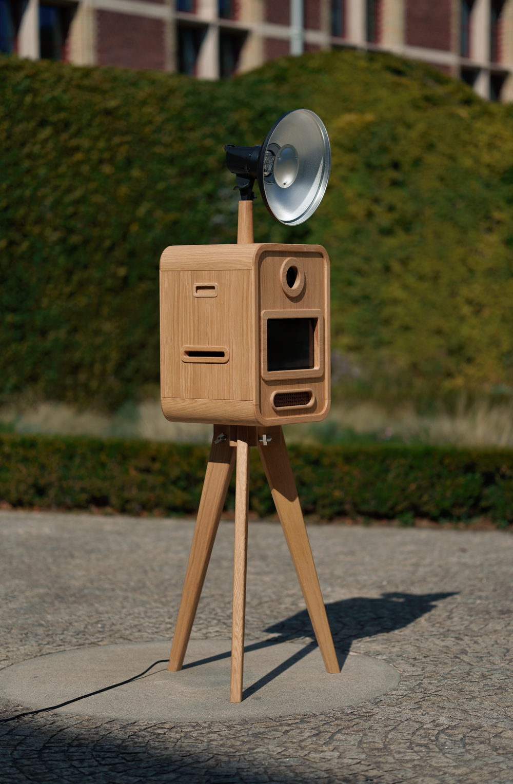 Vintage-style wooden photo booth on tripod with silver flash reflector outdoors on a sunny cobblestone plaza.
