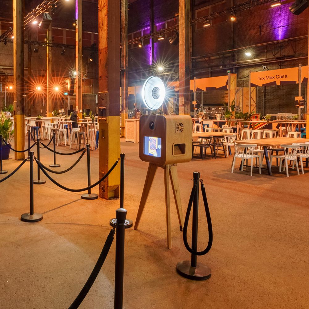 Wooden photo booth on tripod with ring light behind velvet rope barriers in large event hall.