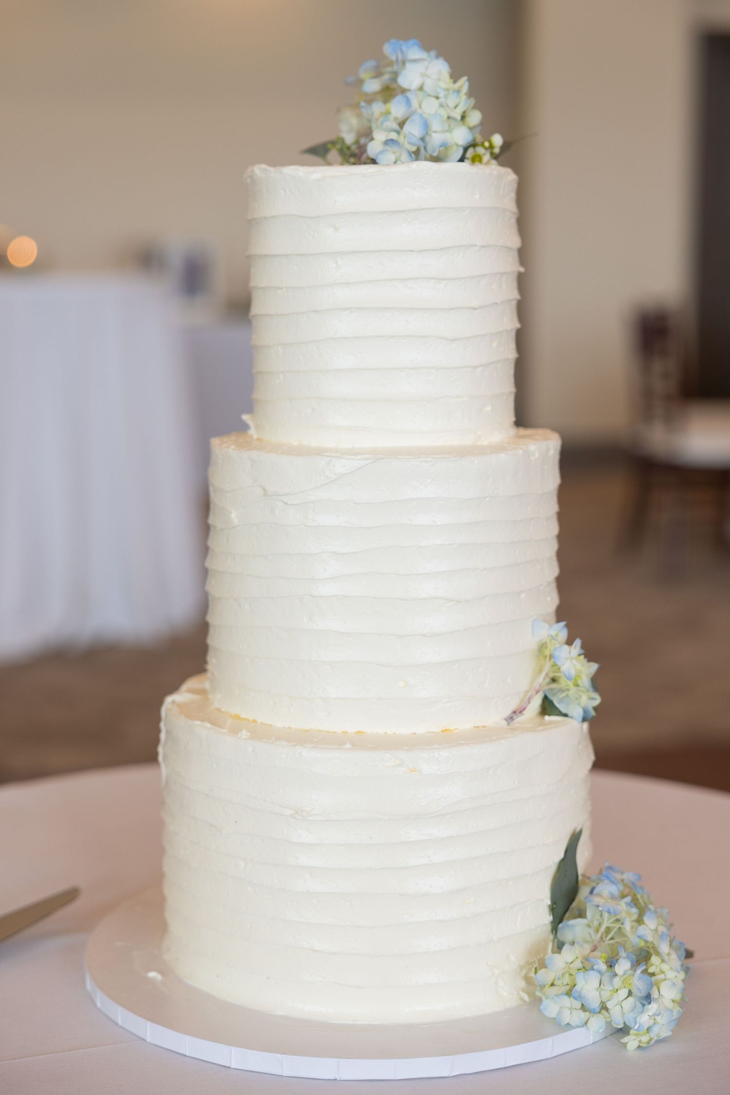 Three-tier white wedding cake with textured horizontal lines and light blue hydrangea flower accents captured by Rachel Martinez Photography