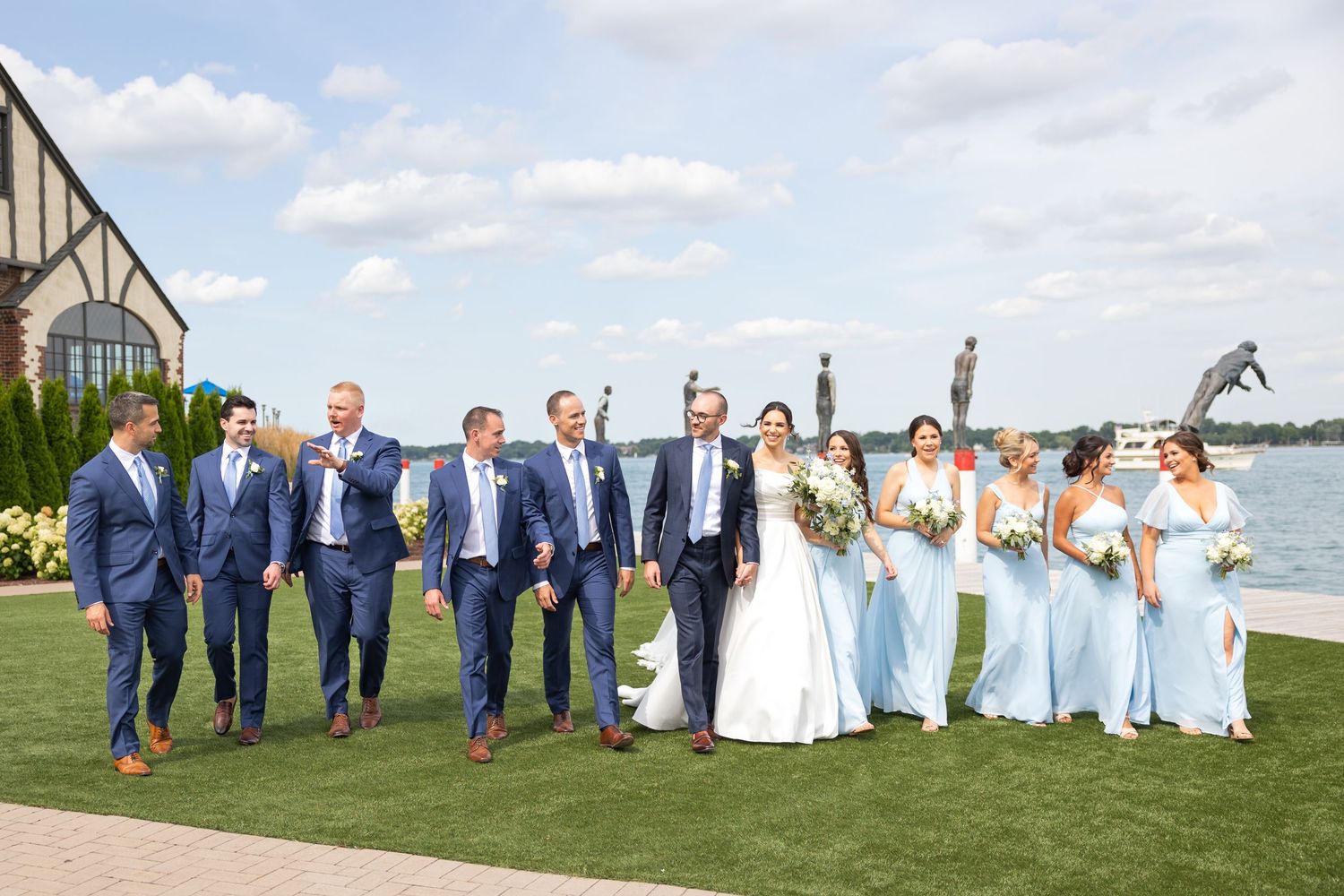 Wedding party in navy suits and light blue dresses pose together on a waterfront lawn on a sunny day.