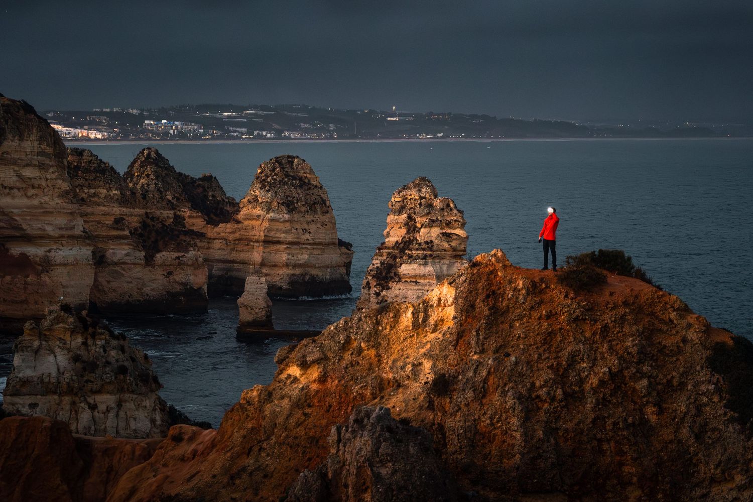 Scenic view of a figure in red jacket standing on rocky cliffs overlooking the ocean at Ponta da Piedade in Lagos, Portugal.