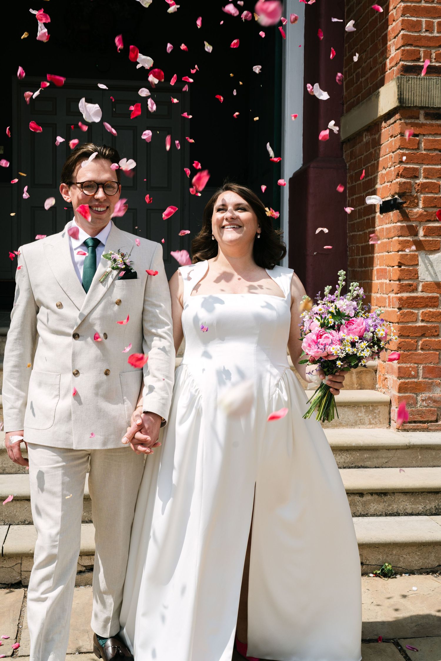 A joyful wedding couple stands on brick steps while pink and white rose petals shower down around them.