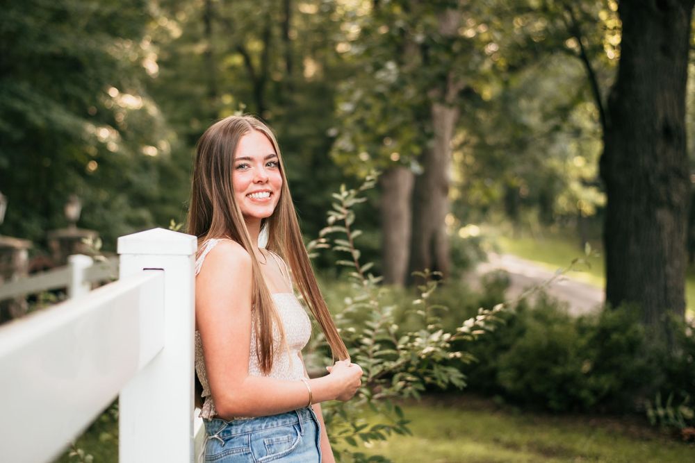Portrait of a senior girl leaning against a white fence with a tree line in the background.
