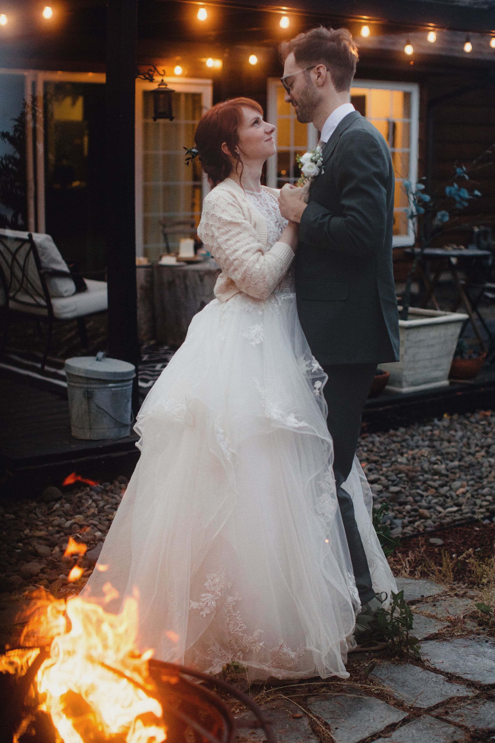 A couple shares a romantic moment by the fire pit at their outdoor evening wedding reception with string lights overhead.