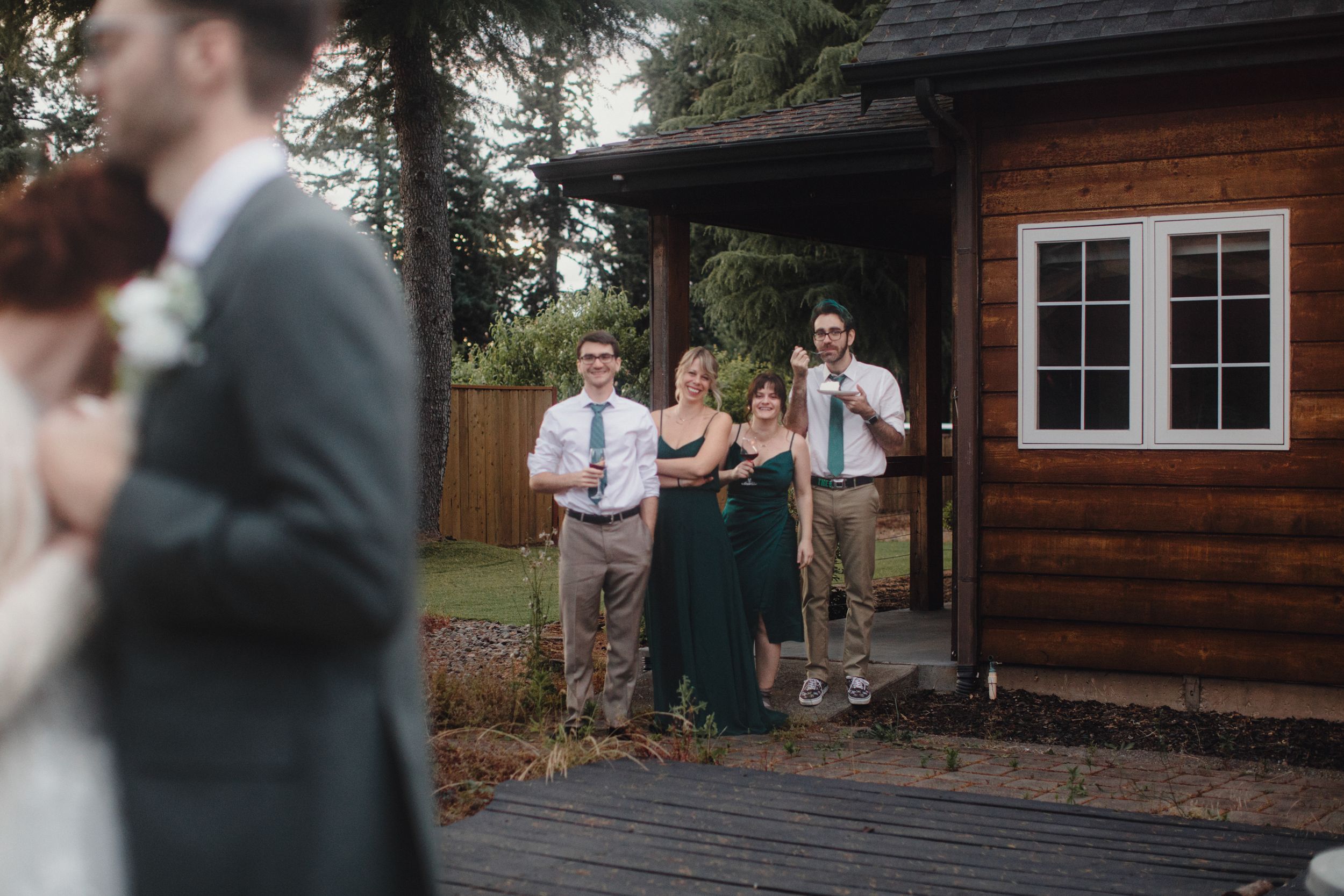 Wedding guests gather outside a rustic wooden cabin venue with dark stained siding and white trim windows.