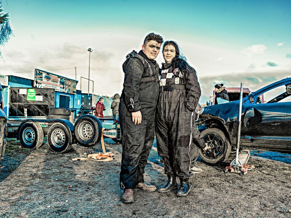 Two banger racers in full racing overalls pose together at a motorsports venue with cars and tyres visible in the background.