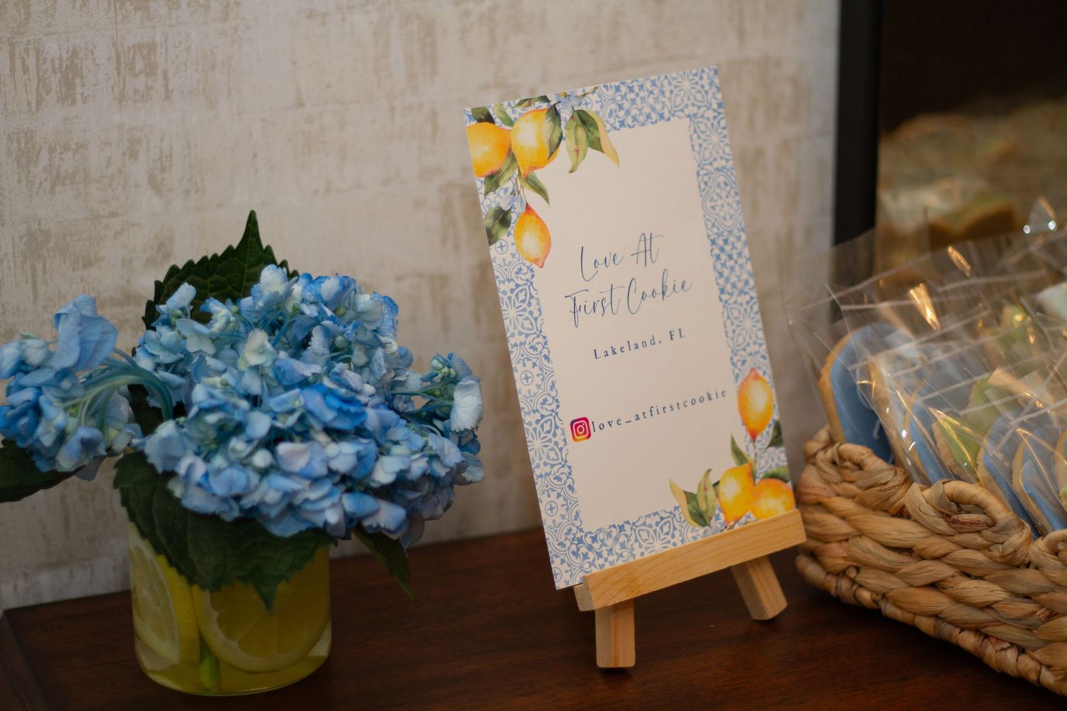 A card on a wooden easel sits next to blue hydrangeas and a gift basket on a table.
