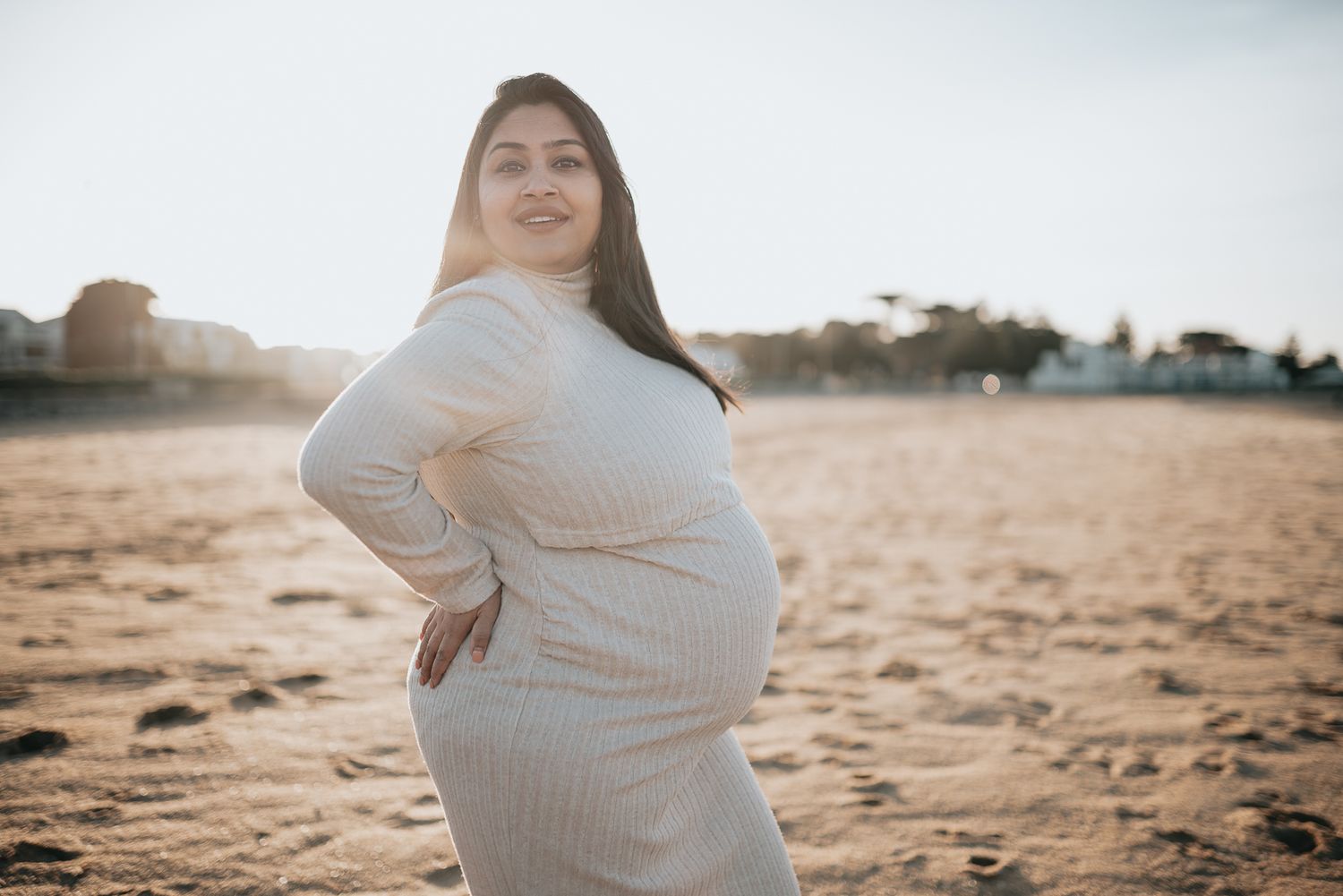 A lady in a white knit dress poses on a sandy beach at sunset with a warm golden glow in the background by Melbourne maternity photographer.