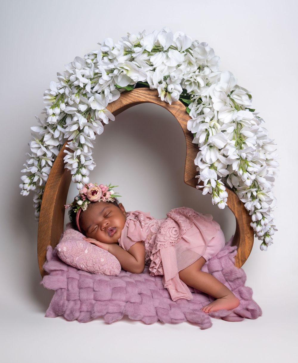 Newborn portrait under floral arch with pink ruffled bedding and white wisteria flowers.