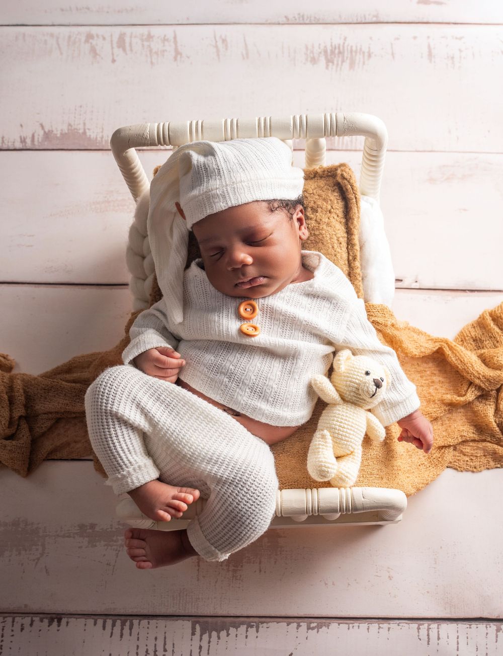 Sleeping baby in white knit outfit lying on wooden surface with plush teddy bear companion.