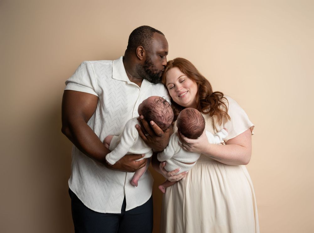 Interracial couple tenderly holding newborn twins against a beige background in a professional studio portrait.