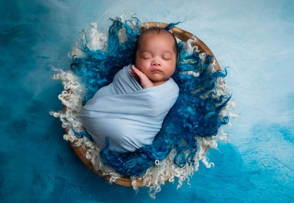 Newborn baby swaddled in blue wrap sleeping peacefully in a wicker basket with blue fluffy decor.