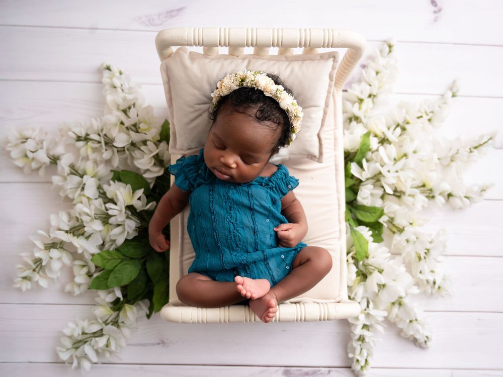 Overhead view of sleeping baby wearing teal outfit surrounded by delicate white flowers on white wooden background.