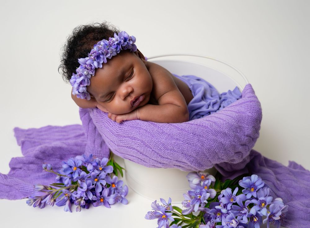 Newborn baby sleeping peacefully in white bowl with purple floral wrap and lavender flowers.