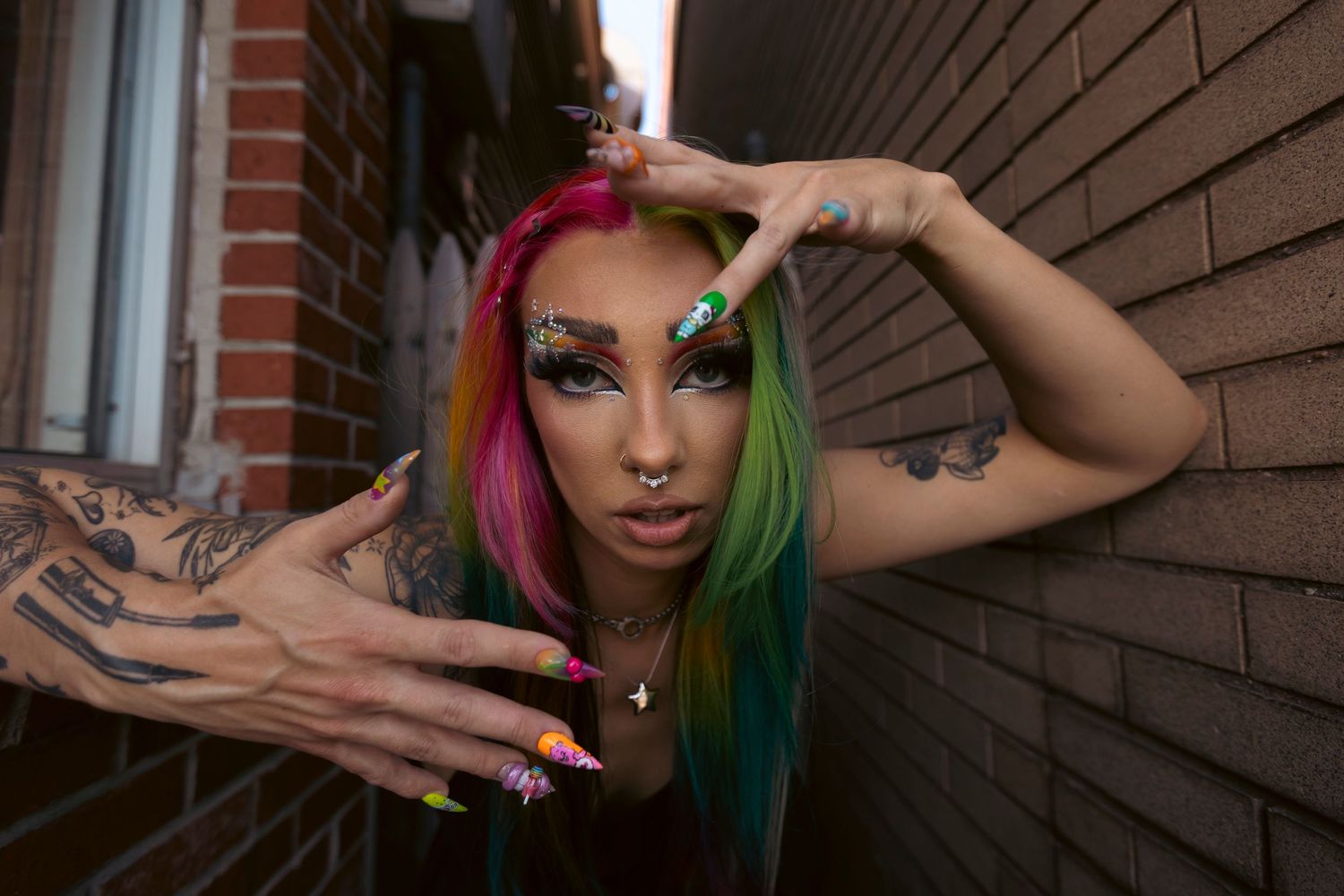 Person with vibrant rainbow-colored hair and artistic makeup poses against a brick wall making dynamic hand gestures.