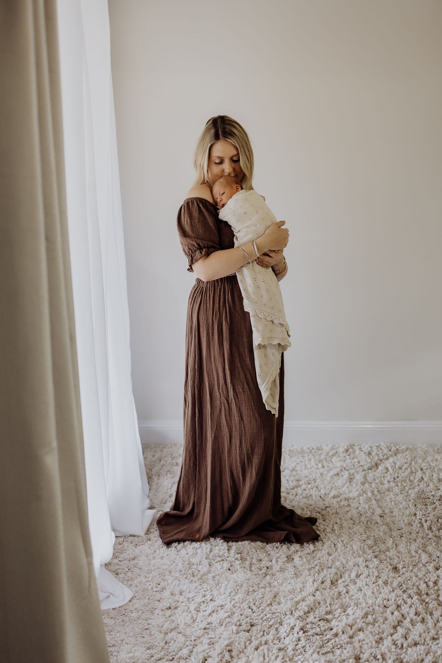 Figure in flowing brown dress holding teddy bear against white wall and textured carpet.