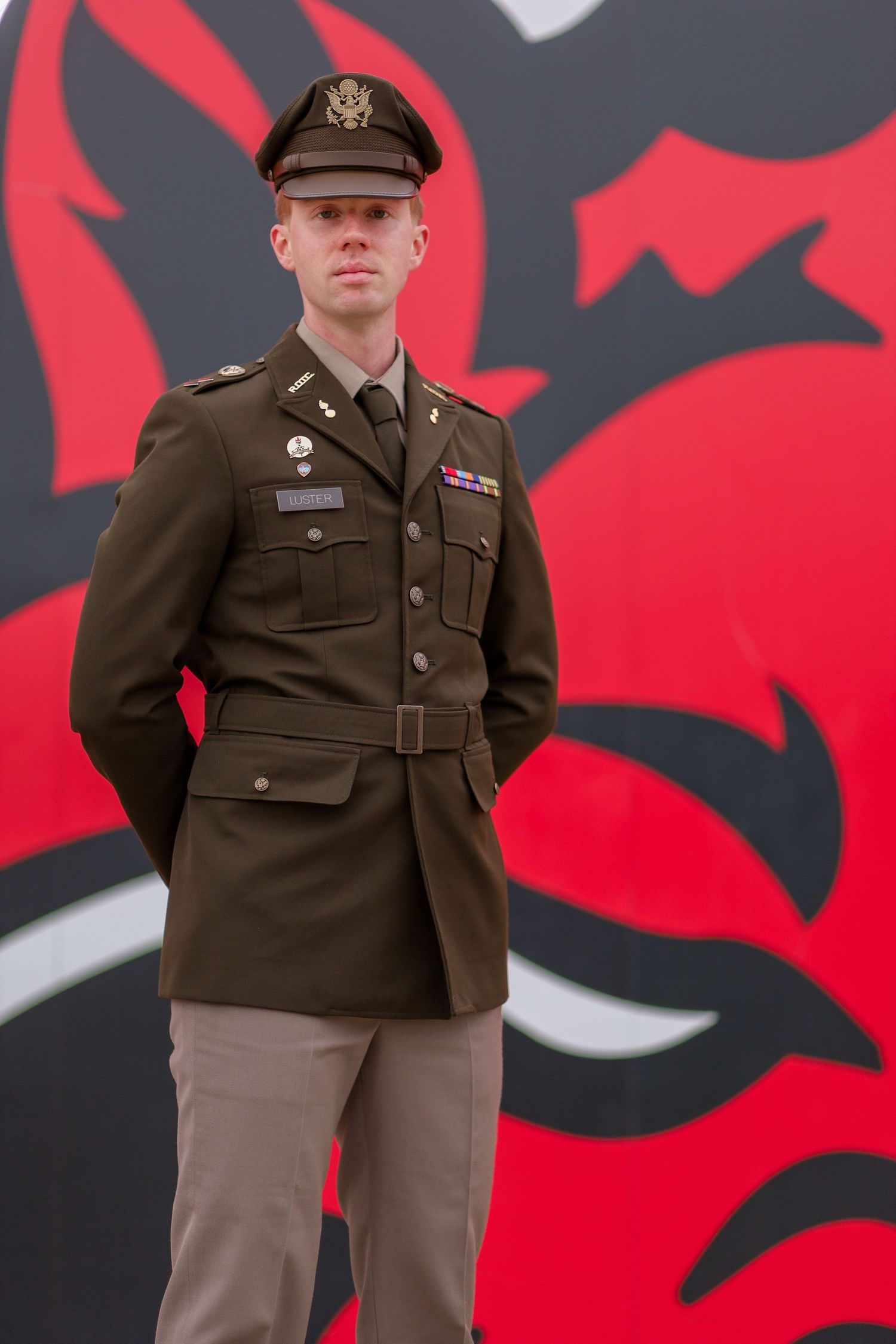 Military service member in dress uniform stands at attention against a red and black painted wall background.