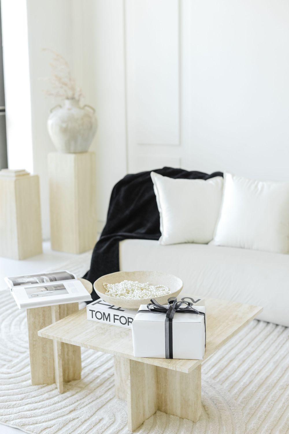 Minimalist white living room with Tom Ford book and gift box on light wooden coffee table against textured carpet.