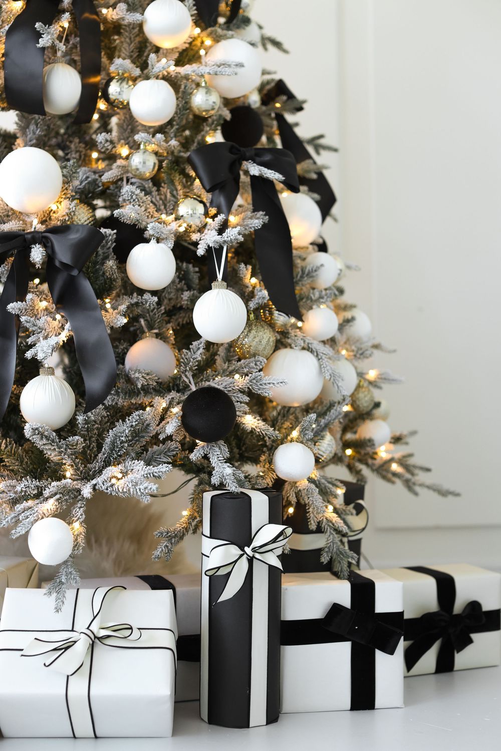 White and black Christmas gifts arranged under an illuminated tree decorated with white ornaments and silver tinsel.