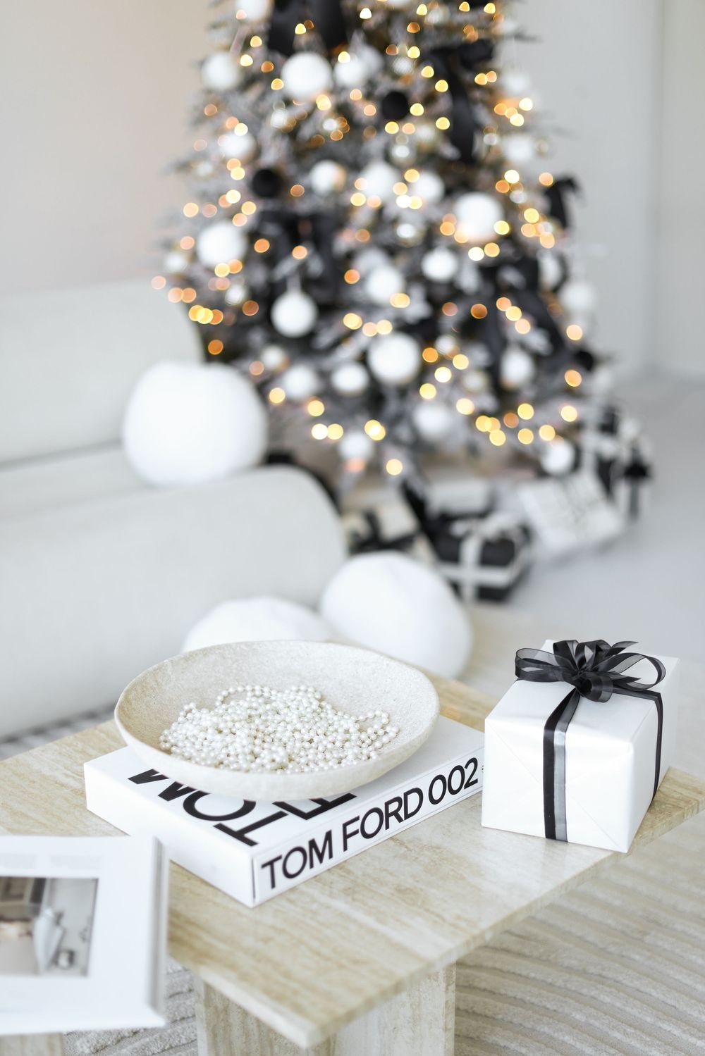 Minimalist Christmas decor with white ornaments, Tom Ford books, and silver beads against a blurred tree with lights.