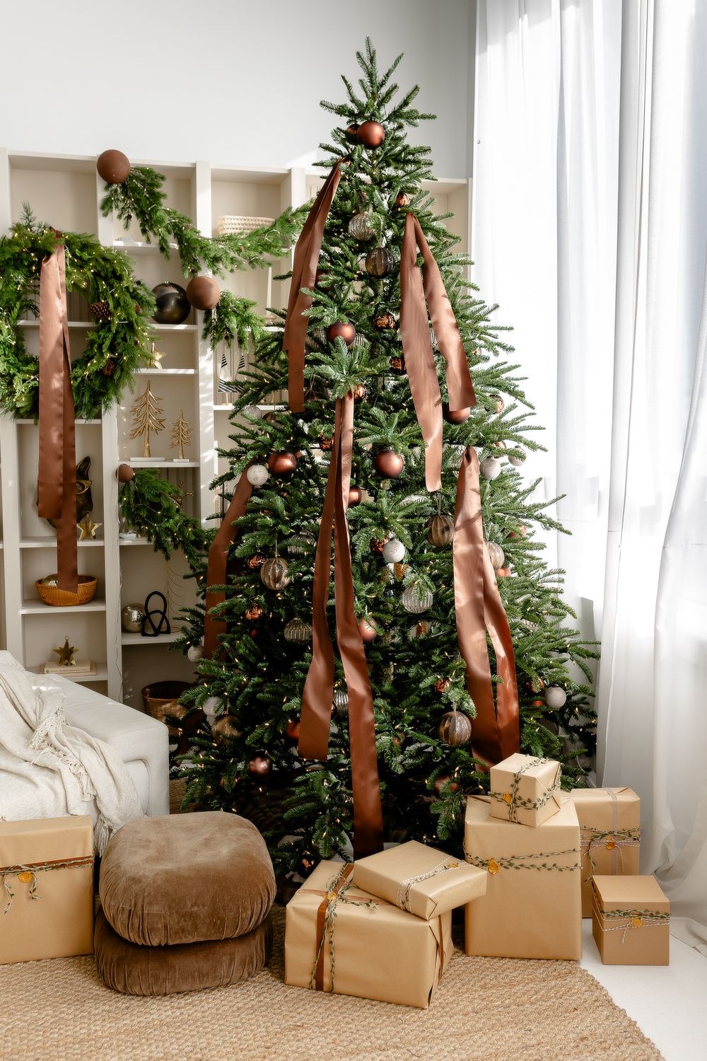 Modern Christmas scene with decorated evergreen tree, brown ribbons and neutral gift boxes near white shelving unit.