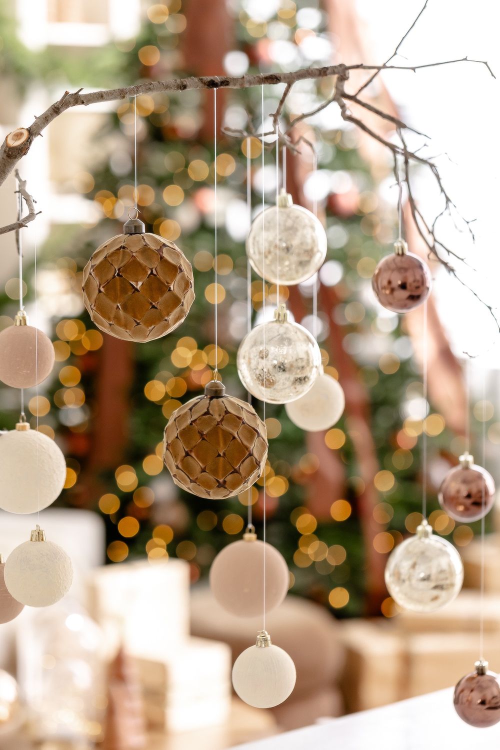 Delicate glass Christmas ornaments in white and rose gold hang from branches with soft bokeh lights in the background.