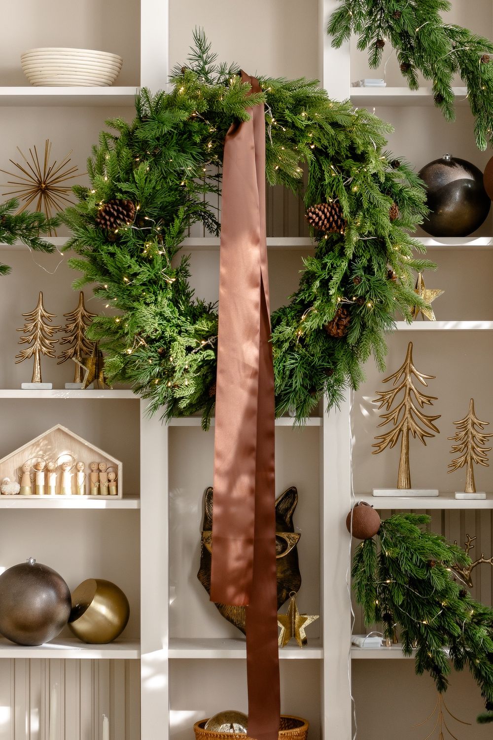 Fresh evergreen Christmas wreath with pinecones hangs on white shelving displaying wooden trees and holiday decor.