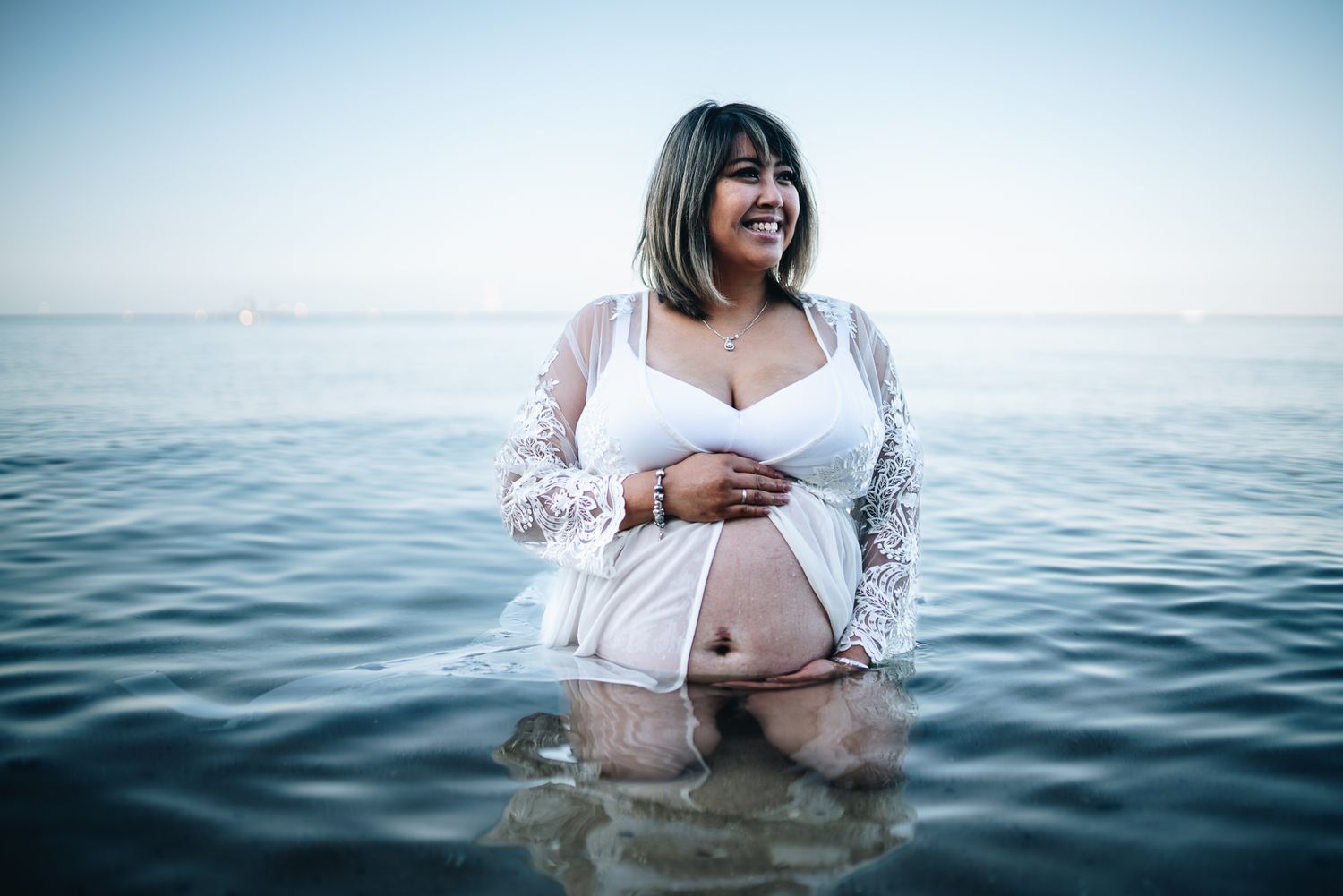 Person wearing white lace top stands waist-deep in ocean water during sunset photoshoot taken by Melbourne maternity photographer.
