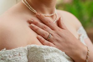 Close-up of hands wearing elegant wedding jewelry and white lace gown showing engagement ring and pearl necklace details.