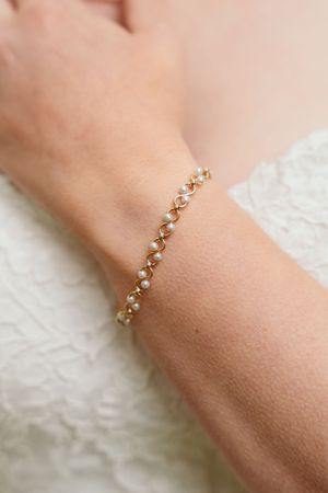 Delicate gold and pearl bracelet worn on wrist against white lace background.