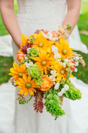 Vibrant orange and white wedding bouquet with sunflowers, green succulents, and coral accents held against a white dress.