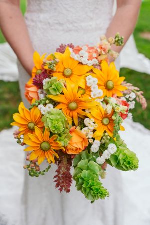 Close up of a vibrant orange and green wedding bouquet featuring daisies, roses and greenery against a white dress background.