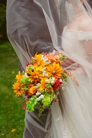 A bright orange and yellow bridal bouquet with daisies and mixed flowers captured through flowing white wedding veil.
