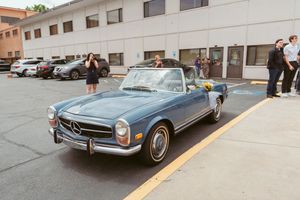 A classic blue Mercedes-Benz 230SL Pagoda convertible parked in a lot beside a commercial building.