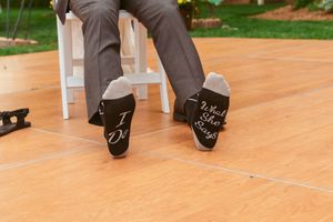 Black dress socks with "I Do" written on the soles are displayed on a wooden deck at a wedding event.
